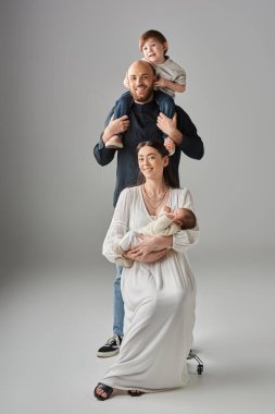 Dört kişilik neşeli aile şık kıyafetler giyiyor ve gri zeminde birlikte poz veriyorlar.