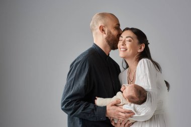 Gülümseyen güzel anne yeni doğmuş bebeği kucağında tutarken kocası onun alnını öpüyor, aile kavramı.