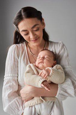 Dikey çekimde güzel, neşeli anne yeni doğmuş bebeğini gri arka planda görüyor.