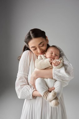 Mutlu annenin uyuyan bebeğini gri arka planda tutuşu, aile kavramı.