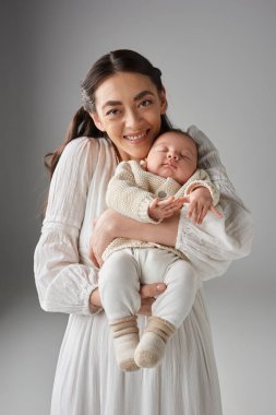 Yeni doğmuş oğluyla, aile konseptiyle poz veren beyaz elbiseli neşeli annenin dikey çekimi.