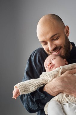 Mutlu sakallı babanın yeni doğmuş bebeğini kucağına alıp neşeyle gülümsediği dikey bir fotoğraf.