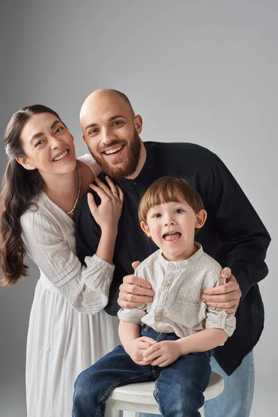 Uzun bir sandalyede oturan küçük oğluna sarılan neşeli modern ebeveynlerin dikey çekimi.