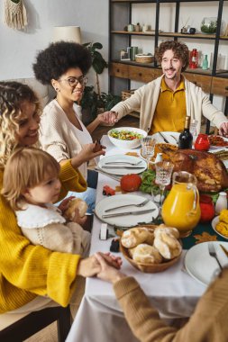 joyful multiracial family holding hands and praying at Thanksgiving table, grateful for dinner clipart