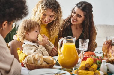 happy multiethnic women looking at toddler girl eating bun during Thanksgiving celebration clipart