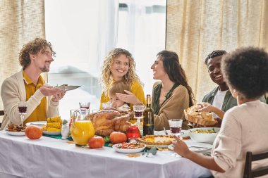 happy multiracial family and friends enjoying meals and drinks while gathering on Thanksgiving day clipart
