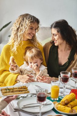 joyful lesbian couple and toddler baby enjoying delicious dinner while gathering on Thanksgiving clipart