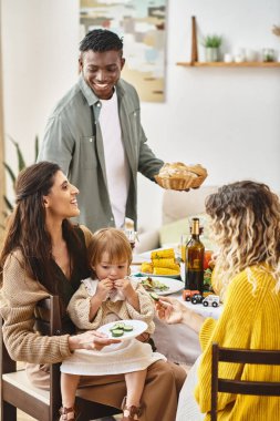 Happy Thanksgiving, curly woman feeding toddler daughter with cucumber near multiethnic friends clipart