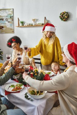Mutlu çoklu etnik arkadaşlar ve Noel Baba şapkalı aileler kadeh tokuşturup mutlu Noeller diler.