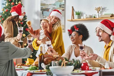 Noel Baba şapkalı, çok kültürlü ve neşeli bir aile Noel masasında şarap bardaklarını tokuşturuyor.