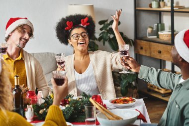 cheerful multicultural relatives in Santa hats raising glasses at festive table on Christmas clipart