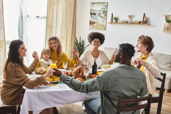 happy multiracial friends and family holding hands and praying at Thanksgiving table, grateful