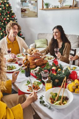 Kırpılmış manzaralı çok ırklı aile bayram yemeğinin tadını çıkarıyor ve arka planda Noel ağacıyla gülümsüyor