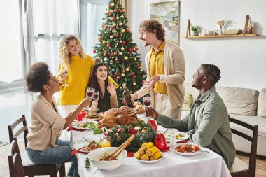 Mutlu aile üyeleri Noel masasında hindi ve şarapla oturup gülüyor ve aktif bir şekilde konuşuyorlar.