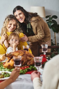 joyful lgbt couple with their daughter in hands smiling cheerfully while sitting at Christmas table clipart