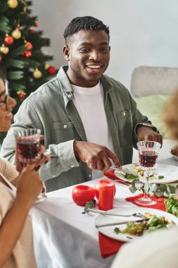 Genç, neşeli Afrikalı Amerikalı adam bayram masasında oturmuş yemek ve şarabın tadını çıkarıyor.