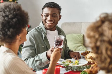 Neşeli Afrikalı Amerikalı adam Noel 'de etrafını akrabalarıyla sarmış şenlik masasında oturuyor.