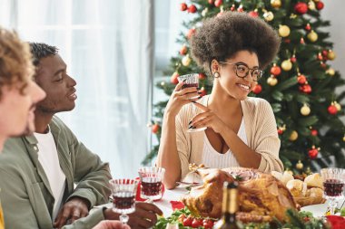 Çok kültürlü akrabalar tatil masasında şarap ve yemekten keyif alıyorlar. Neşeli bir şekilde gülümsüyorlar, Noel.