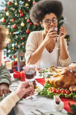 Neşeli Afrikalı Amerikalı kadın akrabalarıyla konuşuyor Noel ağacı arkaplanıyla şarap içiyor