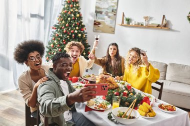 Neşeli çok ırklı aile üyeleri, arka planda Noel ağacıyla şenlik masasında selfie çekiyorlar.