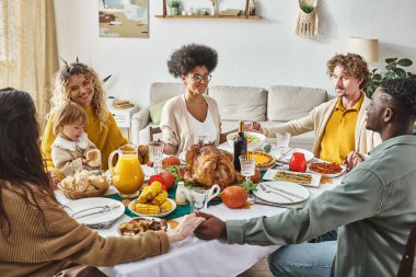 joyful multicultural family members sitting and holding hands praying at festive table, Thanksgiving clipart