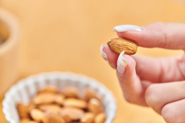 close up view of female hand with almond above bowl with vegetarian food on blurred background clipart