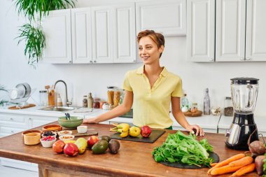 Modern mutfakta çeşitli bitki kaynaklı malzemeler ve elektrikli blender içeren masanın yanında neşeli bir kadın.