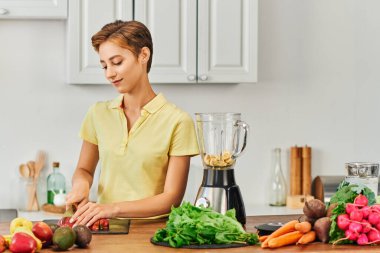 Elektrikli blender yanında elma kesen genç vejetaryen kadın ve sebze ve meyve çeşitleri