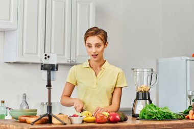 Kadın tripodla akıllı telefonun yanında meyve kesiyor ve evdeki elektrikli blender, bitkisel diyet vlog