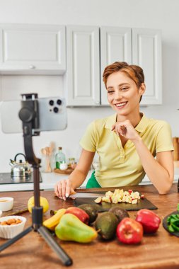 Mutfakta taze meyvelerin yanında tripodla akıllı telefona bakan mutlu vejetaryen kadın.