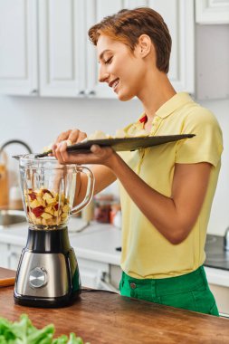 Kesme tahtası olan kadın elektrikli blender 'a doğranmış meyve ekliyor, sağlıklı bitki temelli diyet.