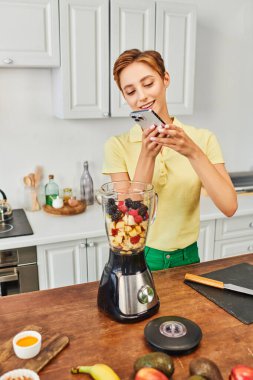 Elektrikli blenderin fotoğrafını çeken neşeli kadın mutfakta taze doğranmış meyveler, bitki temelli diyet.