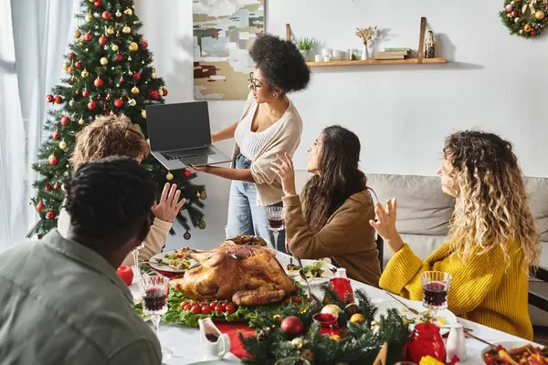 multicultural family gathered at holiday feast waving and gesturing at laptop camera, Christmas