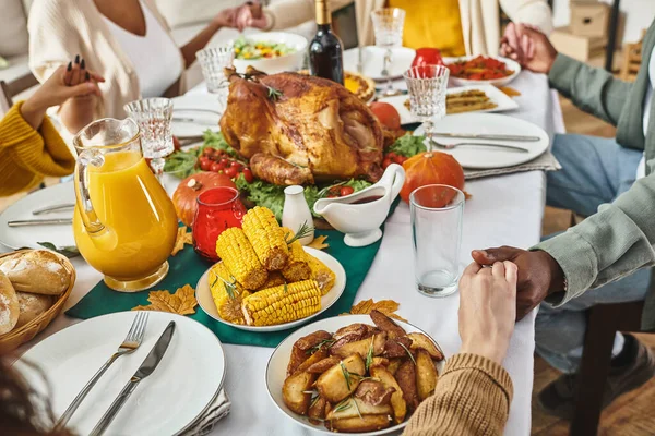 cropped view multiethnic family praying together at festive table with turkey and wine, Thanksgiving