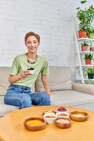 cheerful woman with ripe blackberries near assortment of plant-based food on table in living room