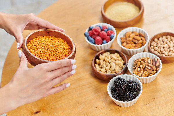 cropped woman with bowl of lentils near table with nuts and fresh berries, plant-based diets concept