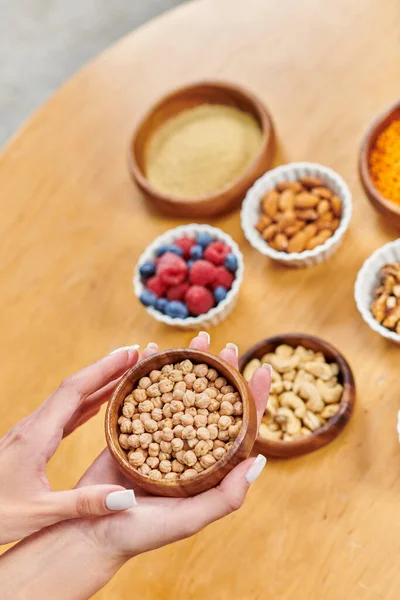 female hands with bowl of chickpeas above plant-based food on blurred table, vegetarian diet