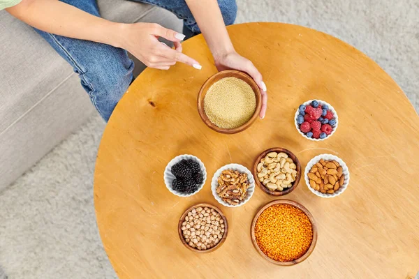 top view of vegetarian woman pointing at bowl with couscous near fresh berries and nuts with legumes