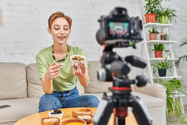 vegetarian woman pointing at bowl of walnuts near plants-based food and blurred digital camera