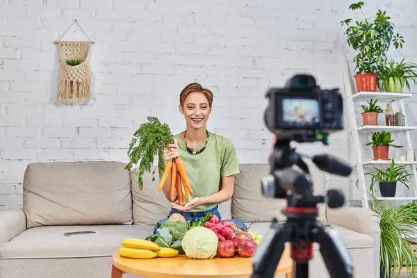 stock image vegetarian video blog, woman with fresh carrots near plant-based food in front of digital camera