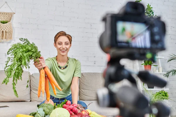 smiling vegetarian woman with fresh carrots talking near fresh vegetables and blurred digital camera
