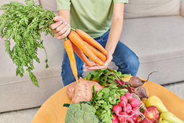cropped view of woman holding fresh carrots above various vegetables and fruits, plant-based diets