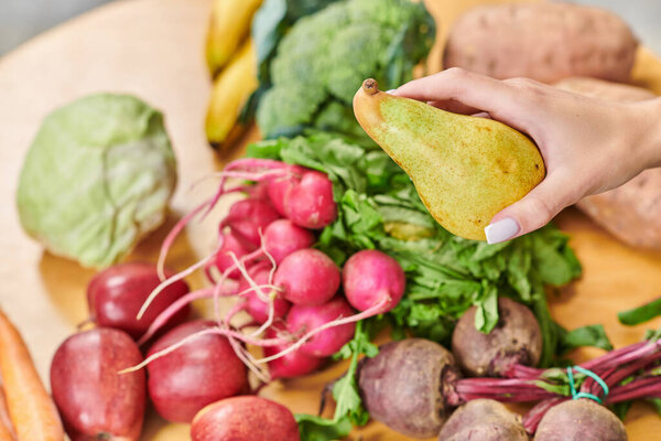 cropped view of vegetarian woman with ripe sweet pier near assortment of fresh fruits and vegetables