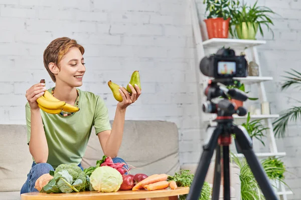 vegetarian woman with ripe bananas smiling during video blog near fresh vegetables and fruits