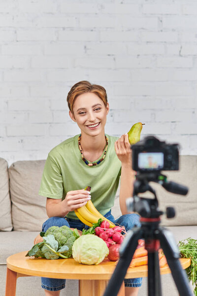 happy woman with ripe bananas near digital camera and fresh fruits and vegetables, vegetarian vlog