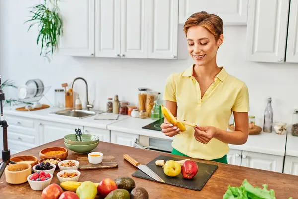 smiley woman peeling ripe banana near fruits and vegetables on table in kitchen, vegetarian concept