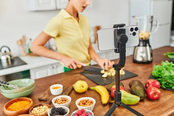 stock image selective focus of smartphone on tripod near vegetarian video blogger cutting fruits in kitchen