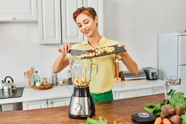 stock image smiley woman adding chopped apple in electric blender near plant origin ingredient, vegetarian diet