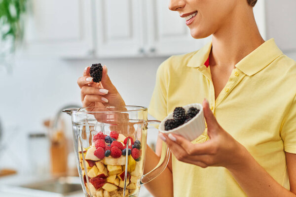 cropped view of smiley vegetarian woman holding ripe blackberries near blender with chopped fruits