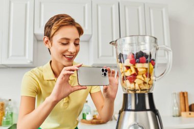 pozitif kadın elektrikli blender ile doğranmış meyve ve lezzetli bitki temelli diyet fotoğrafı çekiyor.
